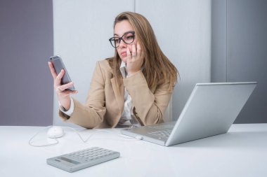 Bej ceketli sarışın kız, çalışma yerinde portalın önünde oturmuş, cep telefonuna sıkılmış bir şekilde bakıyor.