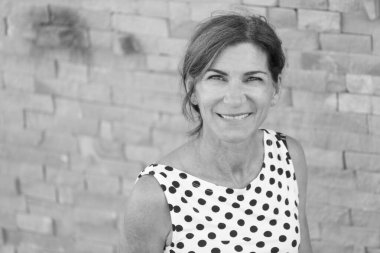 50 yaşında beyaz bir kadının portresi. Puantiyeli bluzlu. Taş bir zemine gülümsüyor.