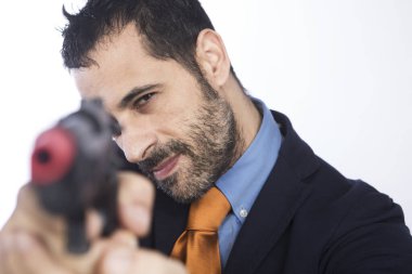 Koyu saçlı, sakallı, turuncu ceketli ve kravatlı adam boş bir silahı işaret ediyor. 