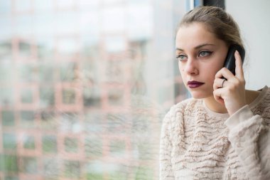 Açık tişörtlü güzel sarışın bir kadının portresi. Telefonda konuşuyor. Büyük bir pencerenin yanında.