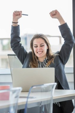 Gözlüklü genç sarışın yönetici iş başarısı için sevinirken masanın önünde dizüstü bilgisayarla oturuyor.