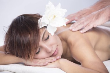 Saçları kısa ve kafasında nilüfer çiçeği olan güzel bir kadın bir spa yatağında dinlenir ve sırt masajına hazırlanır.