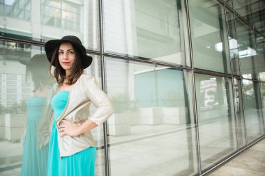 Zümrüt yeşili elbiseli esmer kız ve şapkası modern bir şehir ortamında duvara yaslanırken ayakta duruyor.