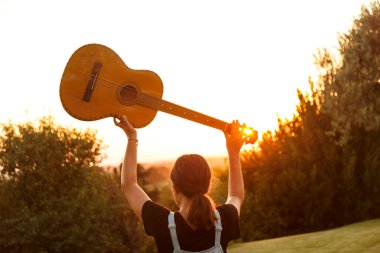 Arkasından gelen kız, gitarını gün batımında doğal bağlamında gökyüzüne kaldırır.