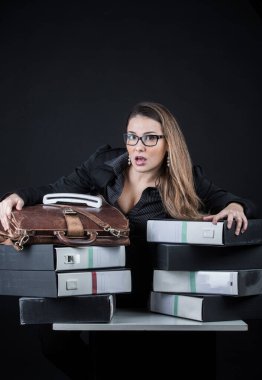 Gözlüklü sarışın sekreter, siyah ve umutsuz giyinmiş ve endişeli. Ofisindeki dağınık dökümanlar arasında, siyah arka planda izole edilmiş.