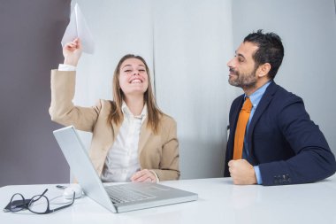 Sivri uçlu takım elbiseli bir adam, masa başında yan yana oturan sarışın bir kızla şakalaşıyor.