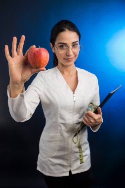 Beyaz önlüklü diyetisyen elinde siyah bir klasör ve mavi bir arkaplanda gururla izole edilmiş bir elma tutuyor.