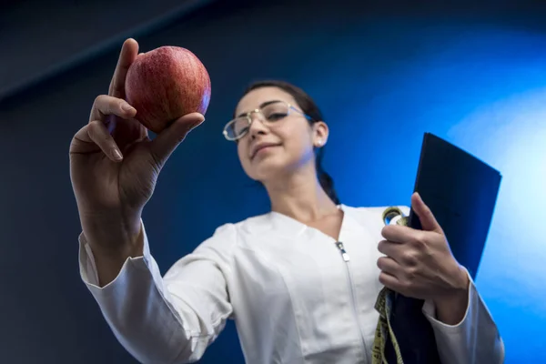 身穿白衣的女孩拿着一个带有测量带的文件夹 上面有一个苹果 表明她的饮食健康而均衡 与蓝色背景隔离 — 图库照片