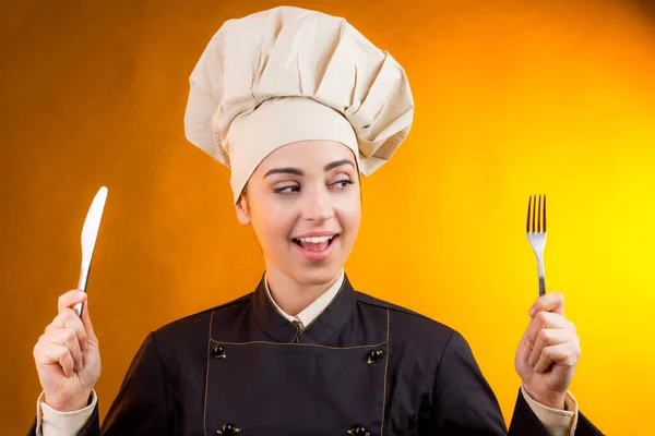 戴着厨师帽微笑的厨师手里拿着一把叉子和一把小刀 用橙色背景隔开 — 图库照片