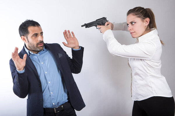 блондинка в белой рубашке направляет пистолет на темноволосого мужчину, одетого в пиджак и рубашку, который поднимает руки, чтобы сдаться, изолированного на белом фоне