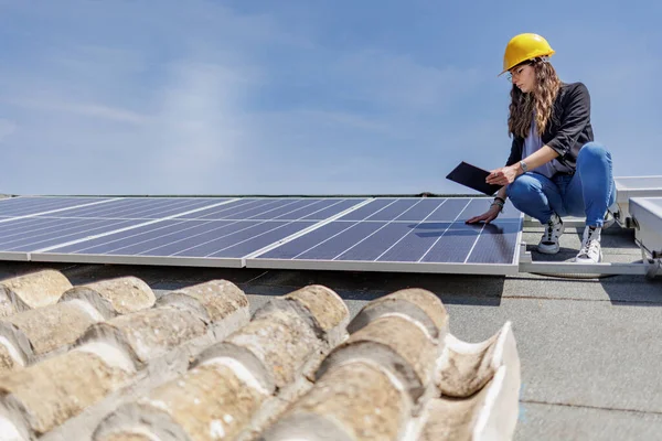 Female engineer in a yellow hard hat checks the condition of a photovoltaic system on the roof of a house with a tablet in her hand