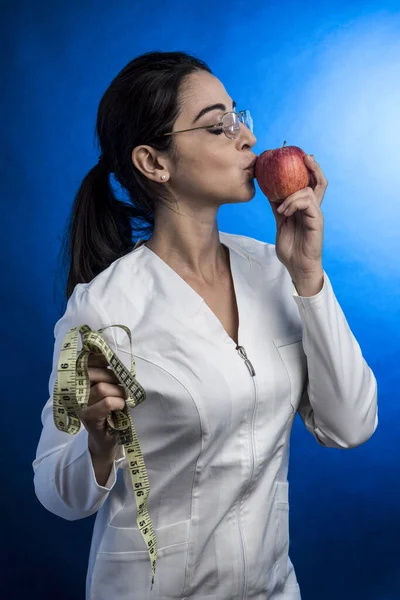 身着白衣 头发蓬松的营养师亲吻着她另一只手拿着的苹果 那只苹果被蓝色的背景隔开了 — 图库照片