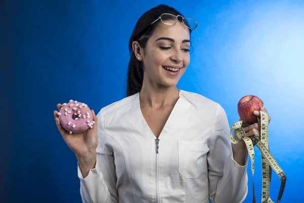 身着实验室外套的营养师 她的头发收集在一只手拿着一个蛋糕 另一只手拿着一个苹果和一个测量身体尺寸的仪器 用蓝色背景隔开 — 图库照片