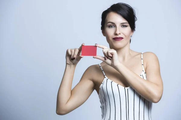 White woman with black hair dressed in a white striped jumpsuit points to a red card, isolated on white background