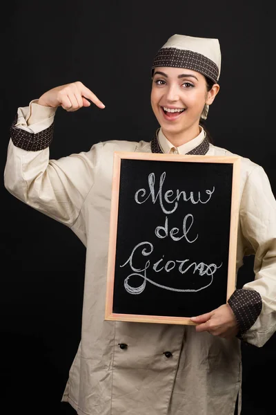 制服姿の笑顔のシェフが イタリア語で メニュー ジョルノ と書かれた黒板を手に持っており オレンジを背景に孤立した 当時のメニュー を意味する — ストック写真
