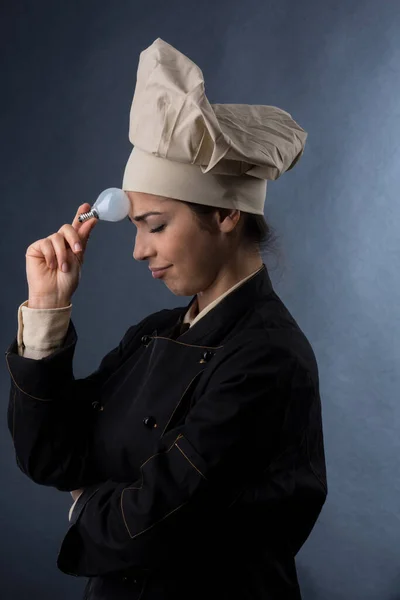 穿着厨师制服的女厨师手里拿着一个灯泡 表示她正在寻找一个创意配方的好主意 这个配方被淡淡的背景隔开了 — 图库照片