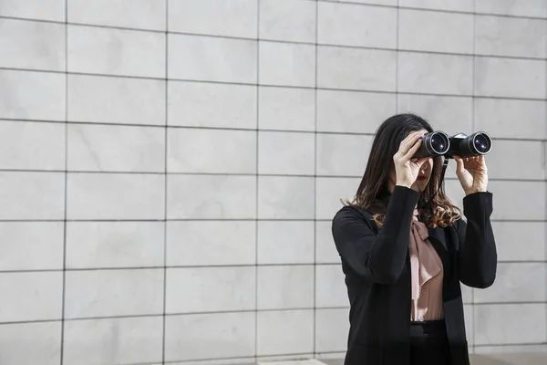 黒のビジネススーツを着たブルネットの女性マネージャーは 白い長方形のタイルの壁の背景に隔離された 離れて見るために双眼鏡を手に持っています — ストック写真