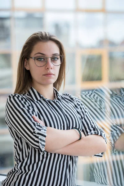 ブロンド女の子でストライプシャツと眼鏡見ます深刻ながら彼女のオフィスに立っていますとともに折り畳まれた腕 — ストック写真