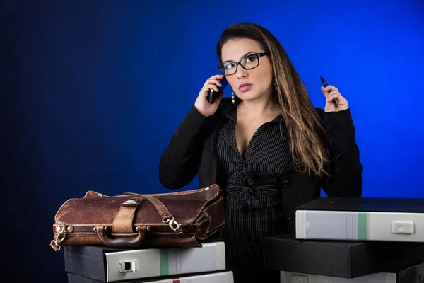 戴眼镜的金发女人经理 身穿黑色西服 坐在办公室里与人通电话 与蓝色背景隔离 — 图库照片