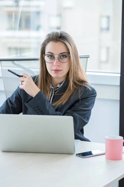 眼鏡をかけた若いブロンドのマネージャーは彼女の仕事の椅子に座っていますノートパソコンと机の前で真剣に見て — ストック写真