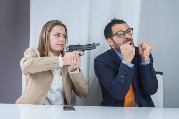 两个同事坐在办公室里 愤怒的秘书用枪指着她绝望的老板 — 图库照片
