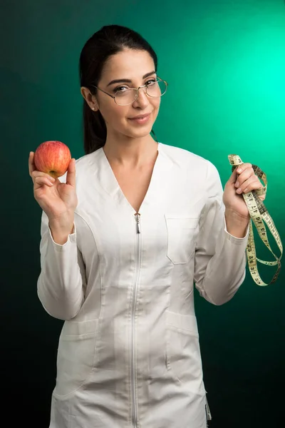 身穿白衣的营养师手里拿着一个测量人体的仪器 还有一个苹果 它骄傲地在绿色的背景下显得孤立无援 — 图库照片