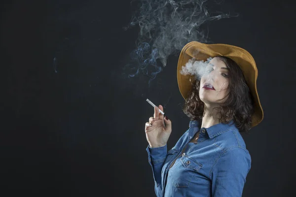 黑发姑娘 穿着牛仔衬衫 头戴牛仔帽 抽着一支香烟 抽着很多烟 背景是灰色的 — 图库照片