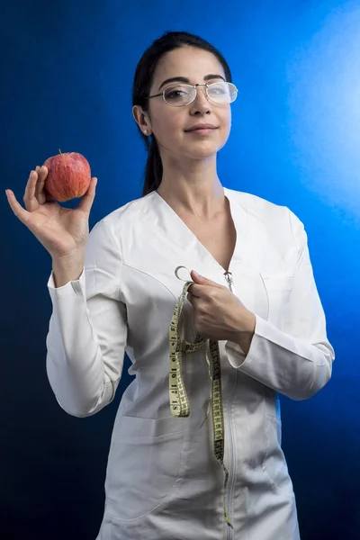 穿着实验室外套的营养师 她的头发收集在一只手拿着一个苹果和一个带尺寸的带子 用蓝色的背景隔开 — 图库照片