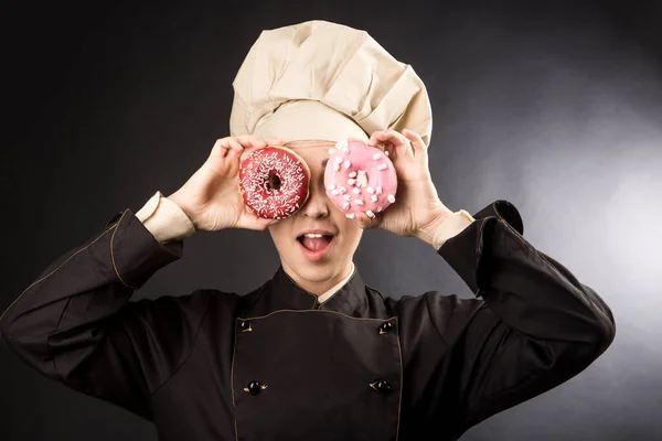 微笑的厨师很好地运用彩色甜甜圈 把它们当作望远镜 用黑色背景隔开 — 图库照片