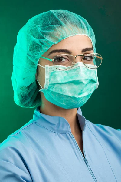 身穿蓝色实验室服 头戴绿色手术室帽 戴着面部外科口罩的医生从他的眼镜中看起来很严肃 与绿色背景隔离 — 图库照片