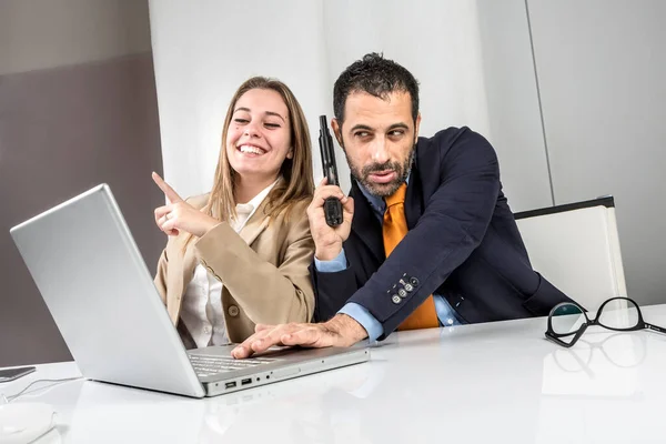 職場に座っている2人の同僚 マネージャーは冗談で銃を手に持っており コンピュータの前で幸せな秘書と密接に協力しています — ストック写真