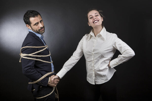 мужчина в пиджаке, связанный веревкой и отчаявшийся перед блондинкой, которая смотрит на него довольной. пара яростно спорит на черном фоне.