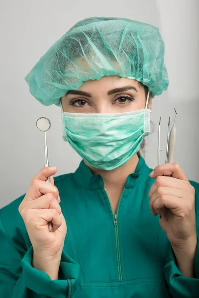 牙科医生 戴着绿色面具 戴着绿色帽子 戴着绿色实验室外套 展示他的工作工具 用浅灰色背景隔开 — 图库照片