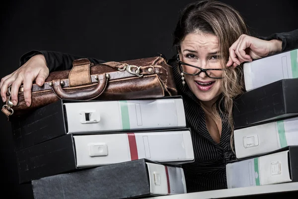 Blonde secretary with eyeglasses, dressed in black and desperate and worried amidst all the messy documents in her office, isolated on black background