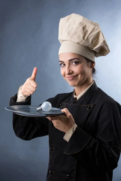 穿着厨师制服的女厨师把灯泡放在盘子里 表示菜谱的意思 然后用大拇指在上面签名 用浅色背景隔开 — 图库照片