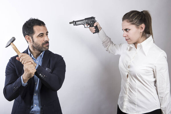блондинка в белой рубашке направляет пистолет на темноволосого мужчину, одетого в пиджак и рубашку, который пытается защитить себя молотком, изолированным на белом фоне