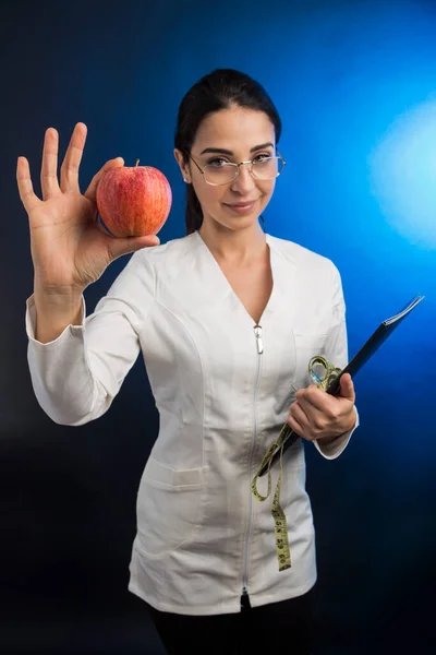 身穿白衣的营养师手里拿着一个黑色的文件夹和一个苹果 在蓝色的背景下骄傲地展示着 — 图库照片