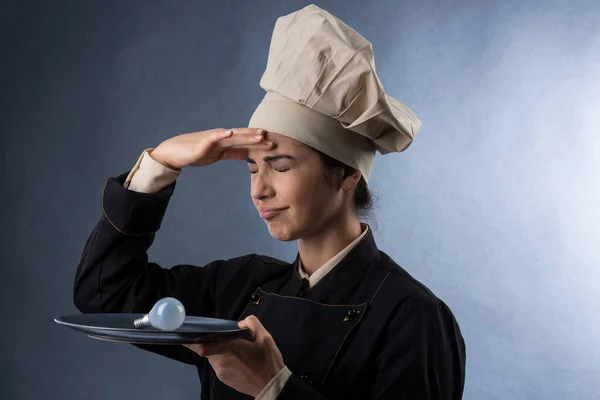 穿着厨师制服的女厨师把灯泡放在盘子里 专心致志地想出一个点子 在明亮的背景下与世隔绝 — 图库照片