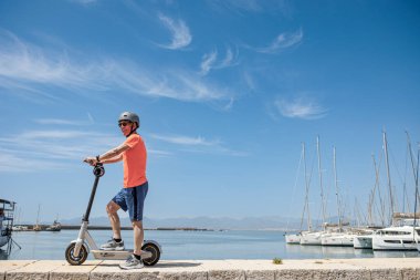 Turuncu formalı ve koruyucu kasklı yaşlı bir adam, marinanın önünde elektrikli scooterına biniyor, arka planda izole edilmiş. 