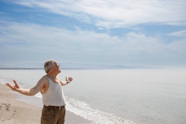 Deniz kenarında askılı tişört giymiş yaşlı bir beyefendi ciddi bir ifadeyle ayakta duruyor.