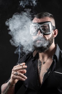 Esmer arka planda dövmeleri olan sigara içen yakışıklı sakallı adamın portresi.