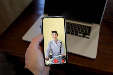 Cep telefonu bağlantısını elinde tutan bir adamın detayları. Ekranda süpermarketten alışveriş yapan bir kadın görülüyor.