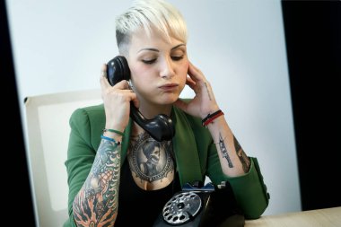 Ofiste yeşil ceketli, sarışın, dövmeli bir kız. Siyah, klasik bir telefonla konuşuyor.