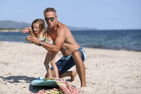 爸爸戴着太阳镜 穿着泳衣 在海滨向女儿传授冲浪的基本知识 — 图库照片