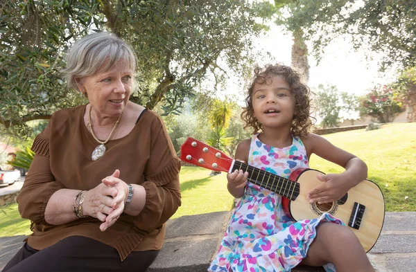 Brazilian granddaughter plays a toy guitar next to her grandmother of European origin sitting in a green park