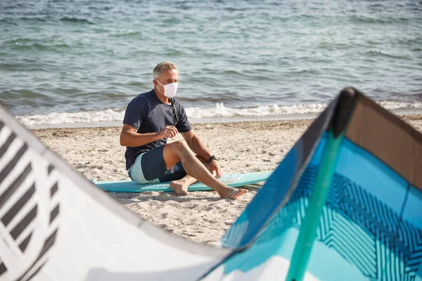 波の帆の横にあるサーフボードに座っているビーチには白いマスクの男が立っています — ストック写真