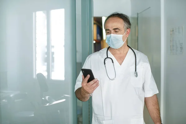Fünfzigjähriger Arzt Weißem Mantel Und Maske Schaut Auf Sein Handy — Stockfoto