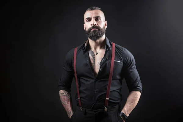Siyah arka planda dövmeleri olan yakışıklı sakallı bir adamın portresi.