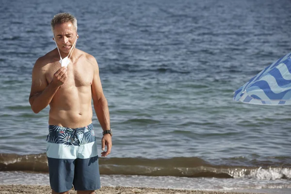 50歳の男性は 保護マスクを脱ぎながら水着でビーチに立っています — ストック写真