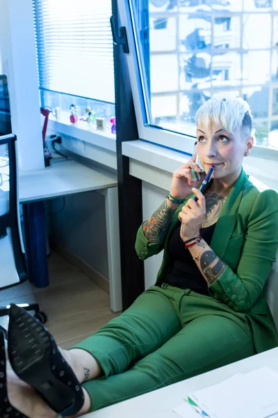 彼女のオフィスの椅子に快適に座って彼女の携帯電話で話して 彼女の体に様々な入れ墨で緑のスーツに身を包んだ — ストック写真